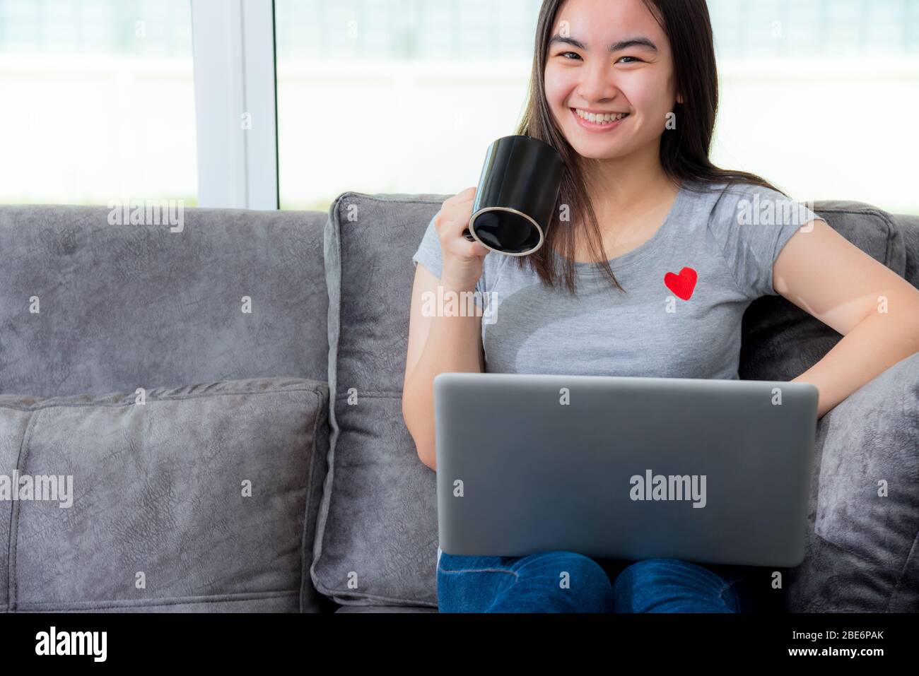 Asiatische Frau mit einer Kaffeetasse Lächeln und während einer Arbeitspause suchen, arbeiten Remote mit einem Laptop auf dem Sofa im Haus, nettes Mädchen Teens glücklich Stockfoto