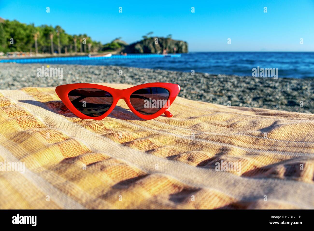 Schwarze Katzenaugen-Sonnenbrille auf einem Handtuch am Strand und Meerblick im Hintergrund. Stockfoto
