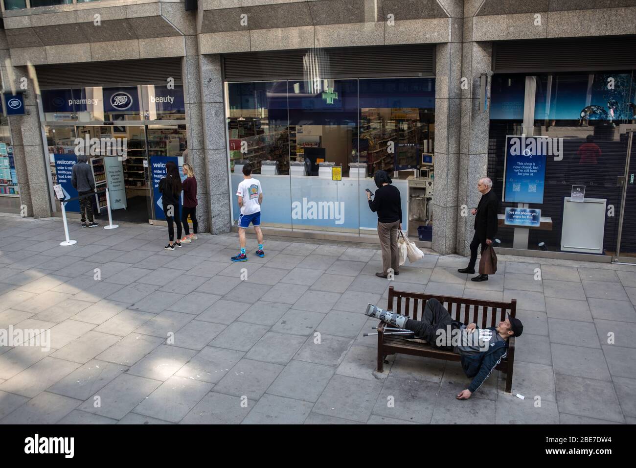 London, Großbritannien. April 2020. Menschen in einer Schlange in sozialer Distanz bei Boots einkaufen, wie ein Obdachloser Mann schläft auf einer Bank während der Sperrung aufgrund von Corona-Virus-Pandemie.Boris Johnson, angekündigt, strenge Lockdown-Maßnahmen drängen Menschen zu Hause bleiben. Rund 78,991 Fälle wurden mit dem Coronavirus (COVID-19) infiziert und 9,875 Todesfälle bestätigt. Das Land befindet sich in der fünften Woche der Blockierung. Das britische Gesundheitsministerium verzeichnete seit Beginn des Ausbruchs insgesamt 84,279 Infektionen und 10,612 Todesfälle. Quelle: SOPA Images Limited/Alamy Live News Stockfoto