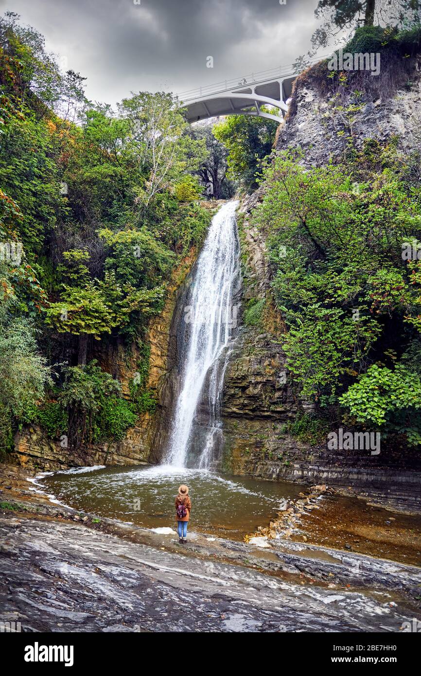 Touristische Frau in Hat in der Nähe des Wasserfalls im Botanischen Garten mit Bäume im Herbst an bedeckt bewölkten Himmel in Tiflis, Georgien Stockfoto