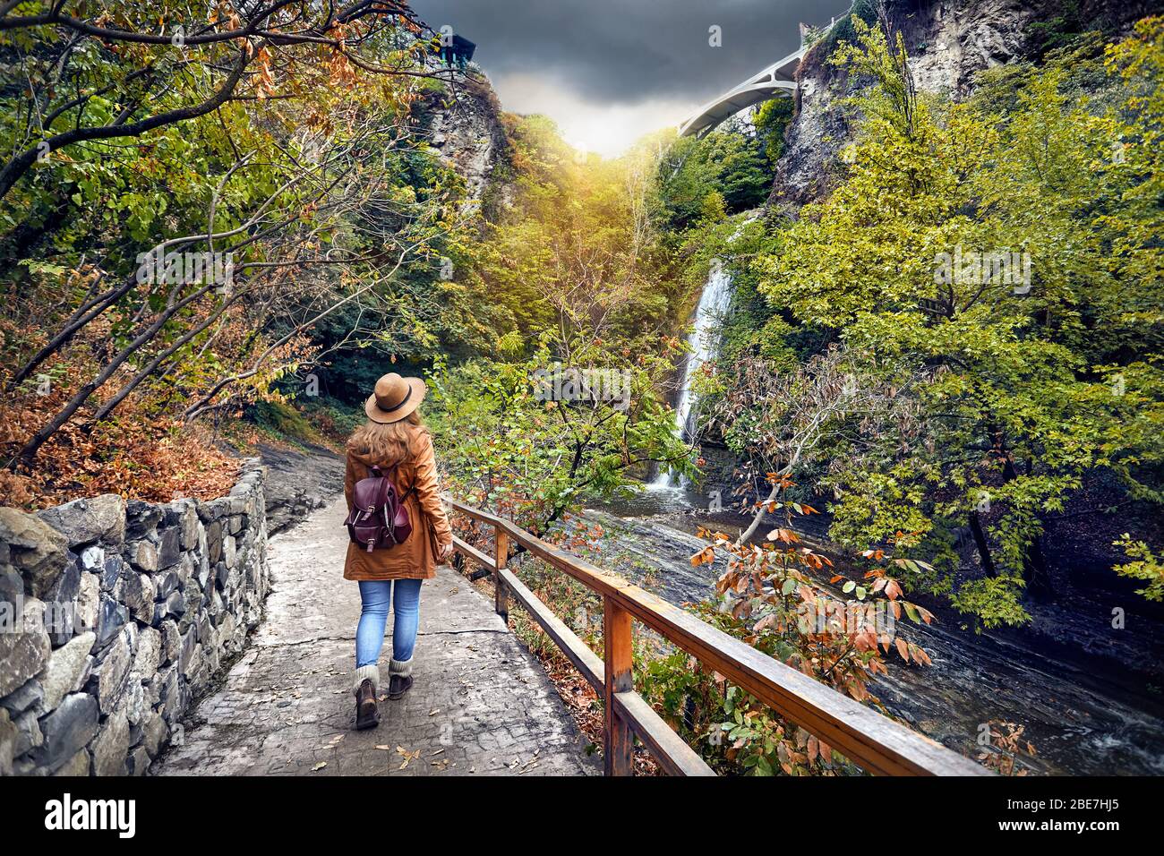 Touristische Frau in Hut gehen zu Wasserfall im Botanischen Garten mit Bäume im Herbst an bedeckt bewölkten Himmel in Tiflis, Georgien Stockfoto