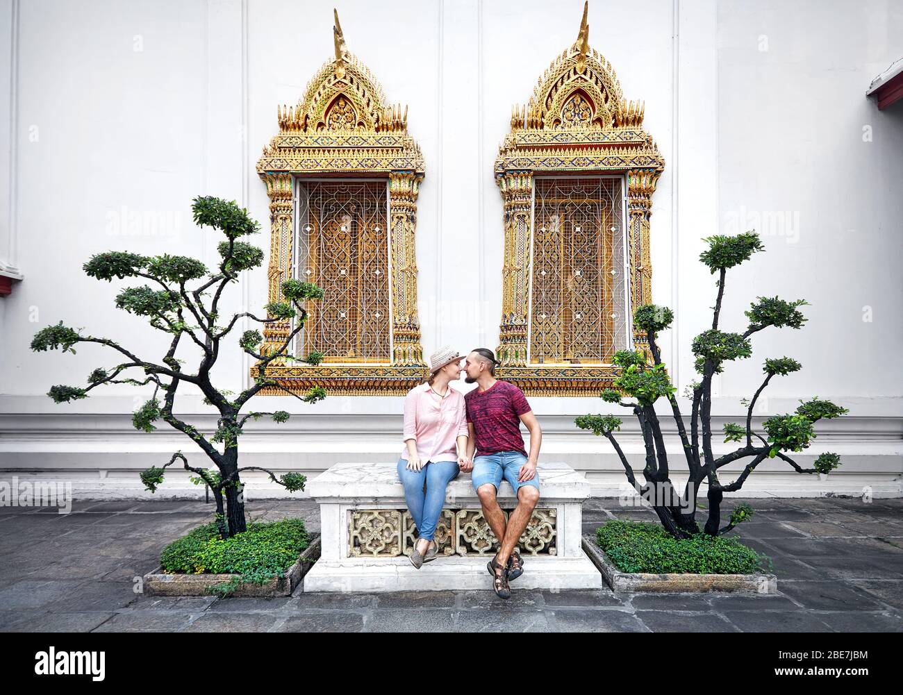 Junges Paar in rote Kleidung auf der Bank in der Nähe von Zierbäume und golden Windows von Wat Pho in Bangkok, Thailand sitzen Stockfoto
