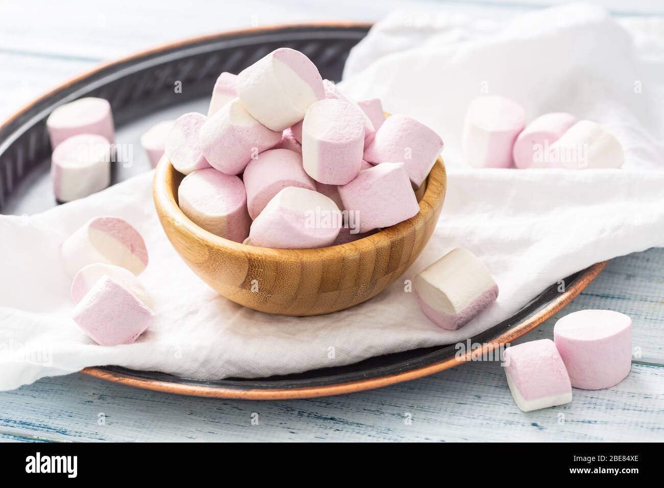 Holzschale voller rosa und weißer Marshmallows mit einigen verstreut auf einem weißen Tischtuch, dunklem Tablett und weißem Holztisch. Stockfoto