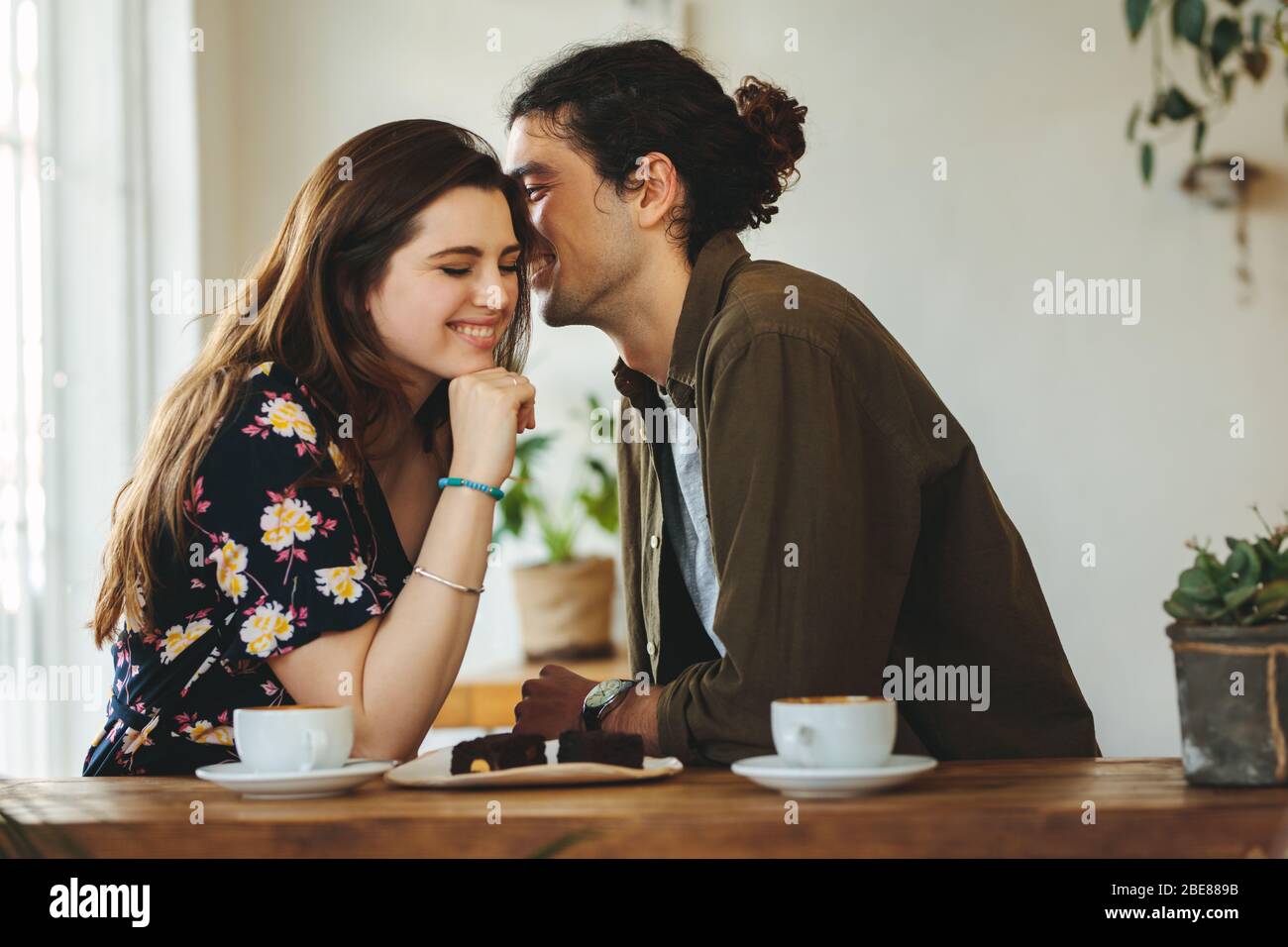 Schönes liebevolles Paar, das in einem Café sitzt und sich in einem Gespräch amüsst. Der Mann flüstert seiner Freundin an einem Kaffeetisch liebevoll ins Ohr. Stockfoto