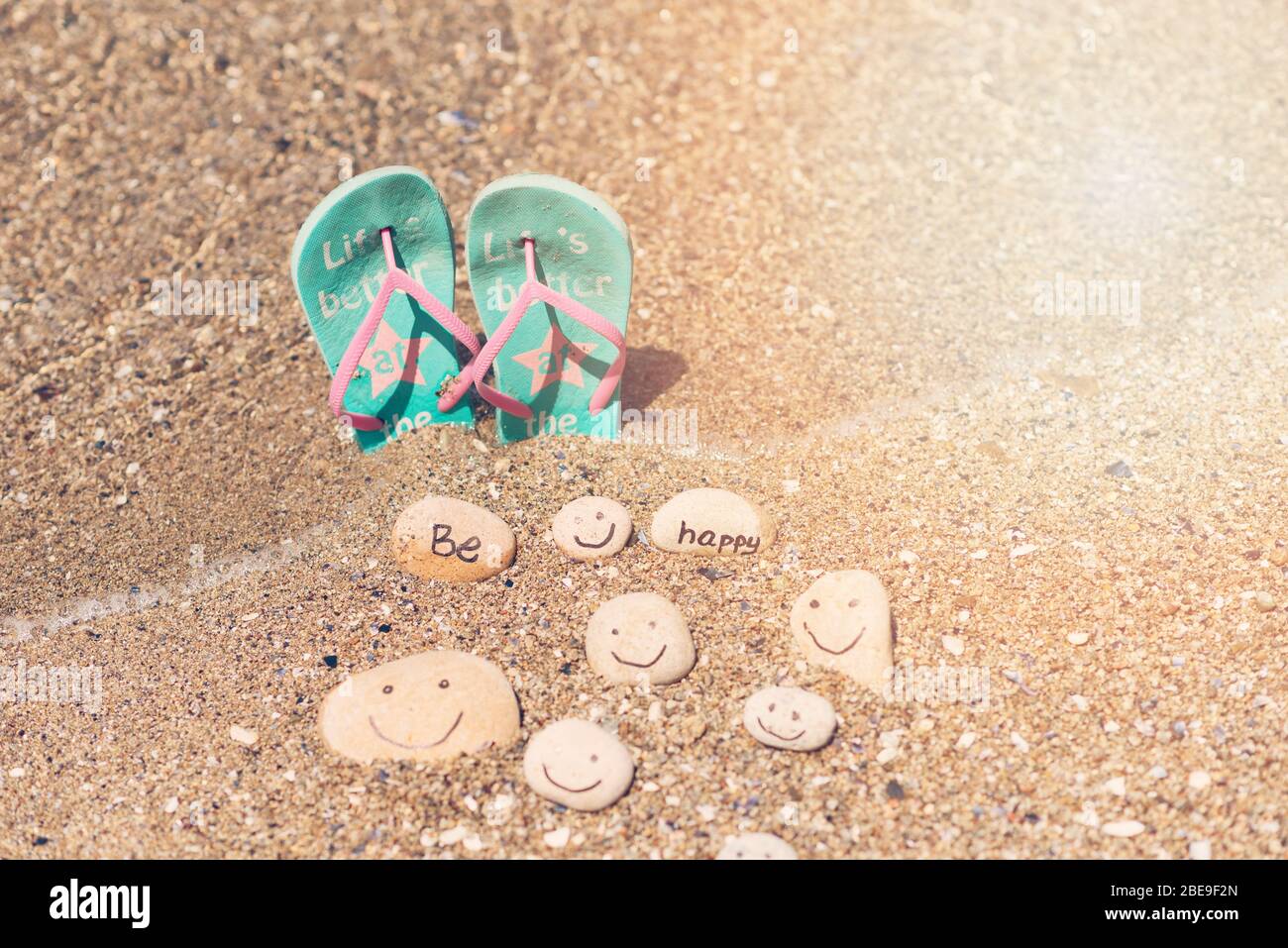Schönes Sonnenlicht mit sauberem Meersand und die Wellen wehen am Strand. Flip Flops und kleine Felsen mit Inschrift, in der Nähe des Wassers. Stockfoto