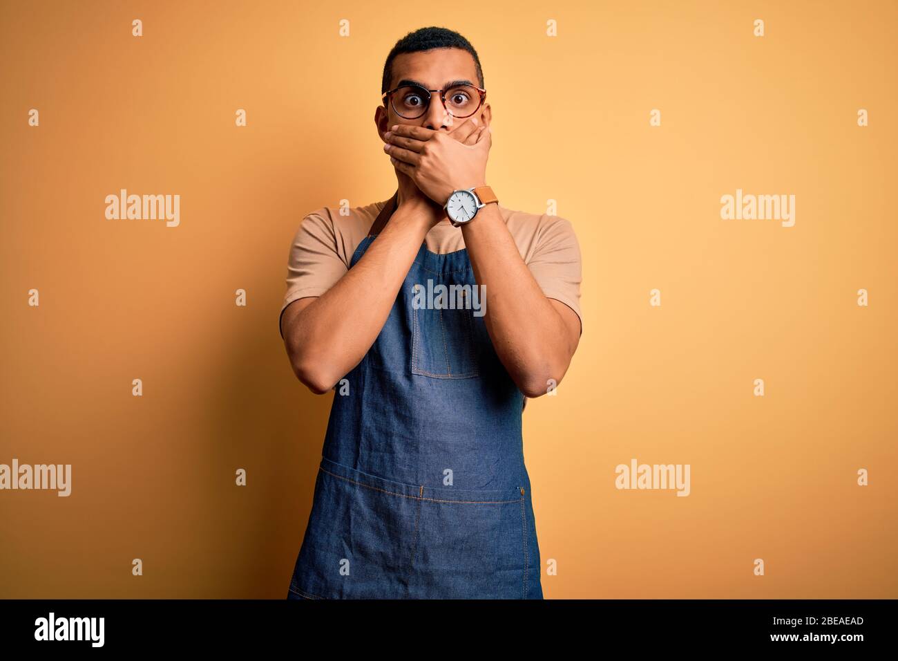 Junge gut aussehende afroamerikanischen Ladenbesitzer Mann trägt Schürze über gelben Hintergrund schockiert Abdeckung Mund mit den Händen für Fehler. Geheimkonzept. Stockfoto