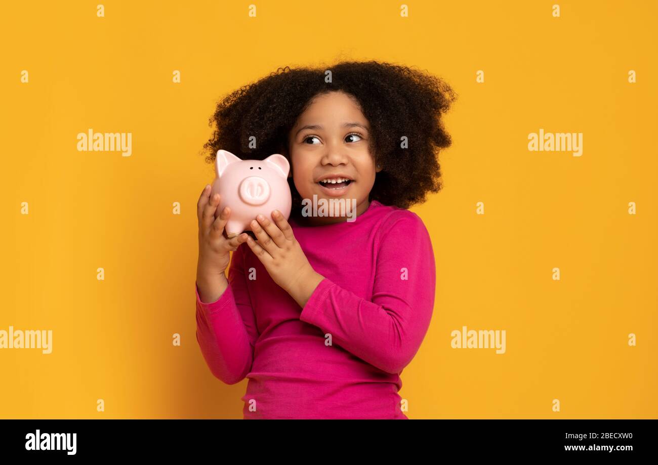 Porträt von entzückenden kleinen schwarzen Mädchen mit ihrem Sparschwein, gelben Hintergrund Stockfoto
