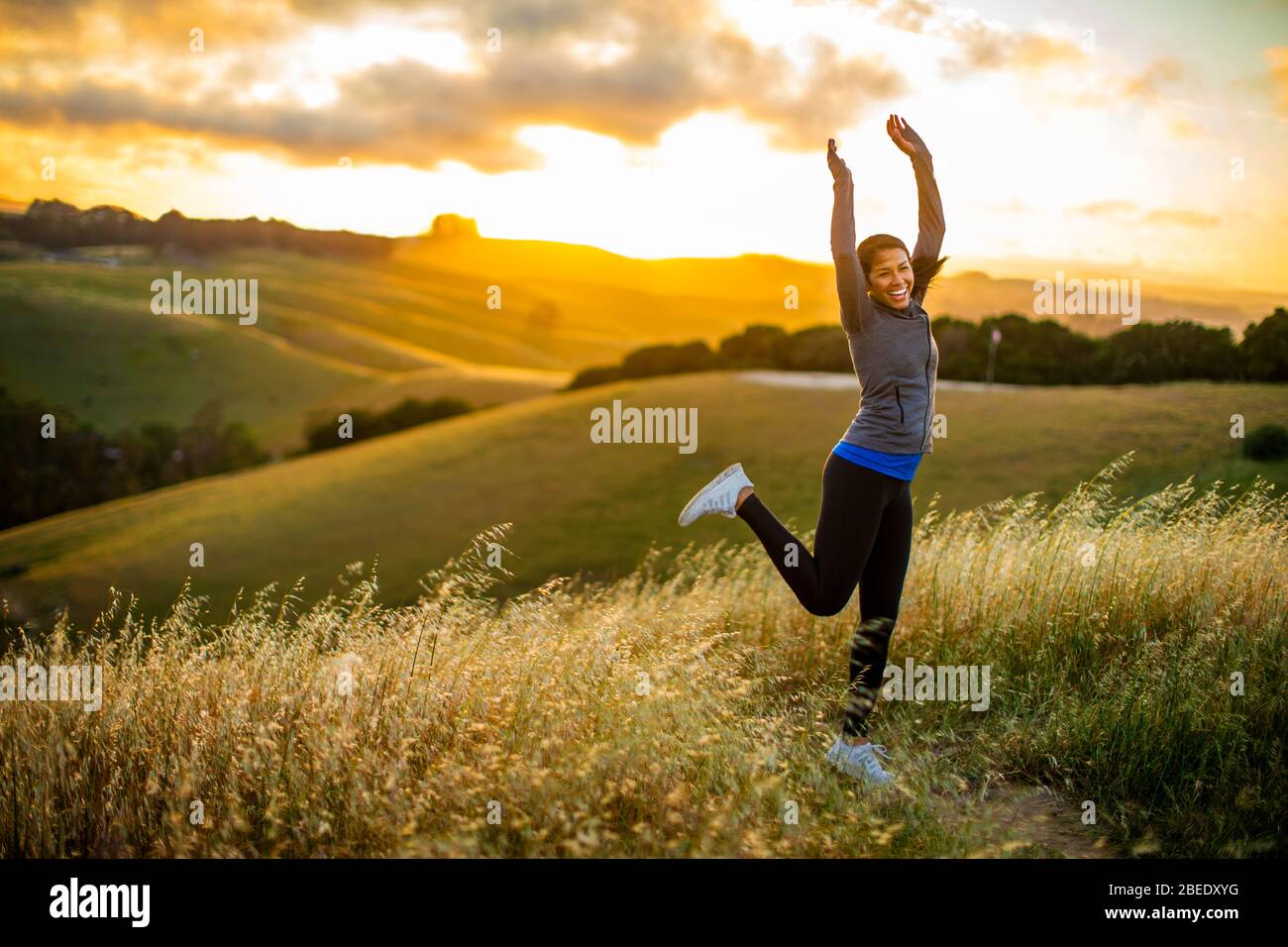 Lächelnde junge Frau springt vor Freude in der Landschaft bei Sonnenuntergang Stockfoto