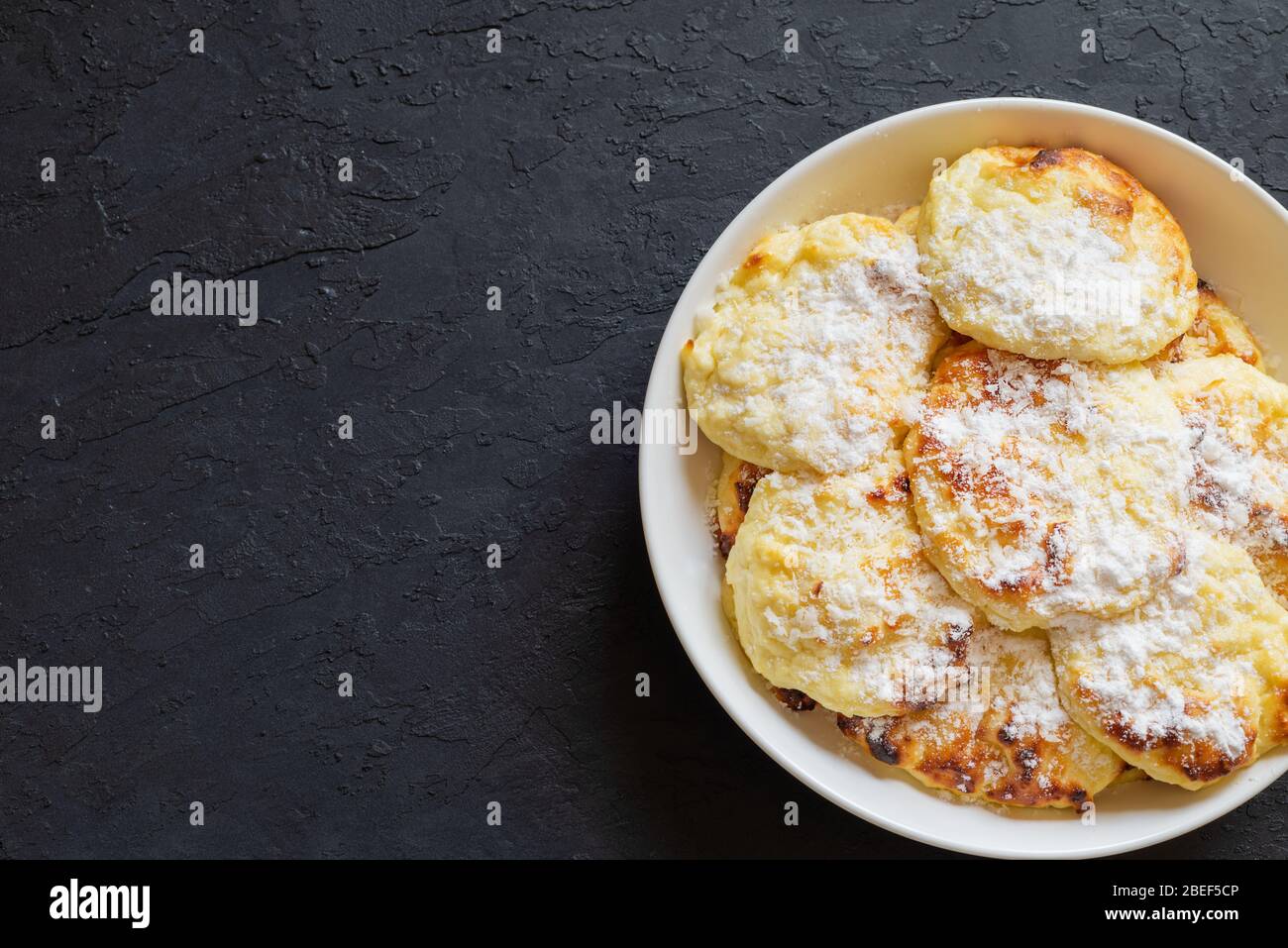 Süße Quark-Pfannkuchen auf dunklem Hintergrund. Pfannkuchen mit Quark auf einem weißen Teller mit Puderzucker bestreut. Kopierbereich. Stockfoto