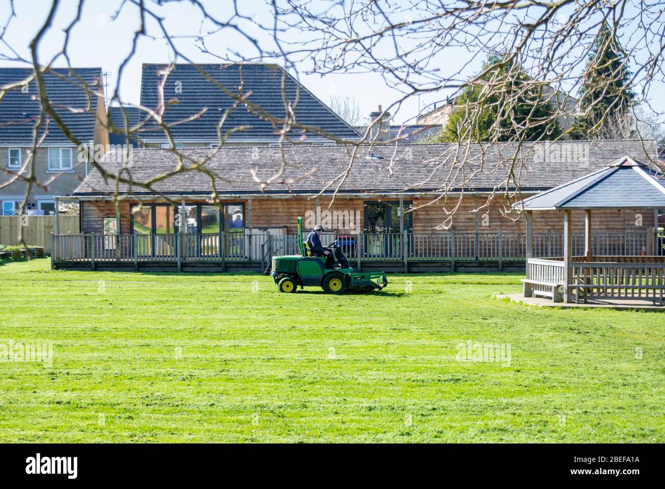 Ein Mann auf einer Fahrt auf Rasenmäher schneidet das Gras eines Grundschulfeldes, während die Kinder nicht in der Schule sind Stockfoto