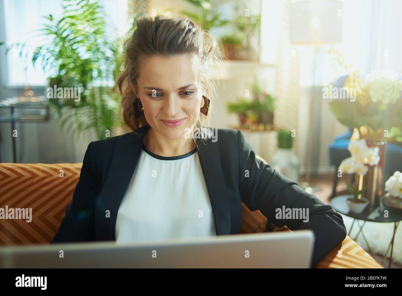 Happy Middle Age Hausfrau in weißer Bluse und schwarze Jacke bei modernen Hause in sonnigen Tag Eingabe Nachricht auf einem Laptop während auf dem Sofa sitzen. Stockfoto