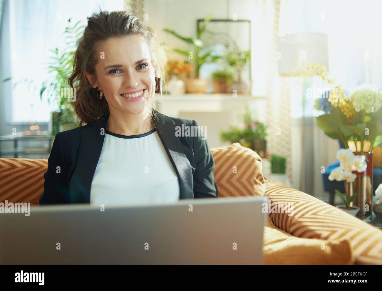 Happy Modern Hausfrau in weißer Bluse und schwarzer Jacke im modernen Wohnzimmer in sonnigen Tag Surfen Web auf einem Laptop, während auf dem Sofa sitzen. Stockfoto