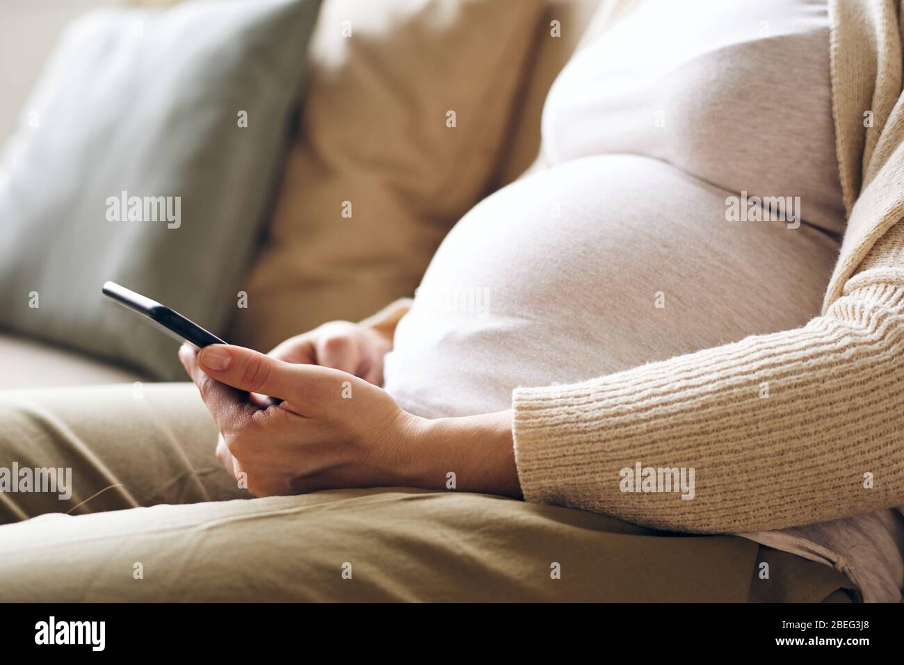 Nahaufnahme einer nicht erkennbaren schwangeren Frau in bequemer Kleidung, die auf dem Sofa sitzt und das Smartphone benutzt Stockfoto