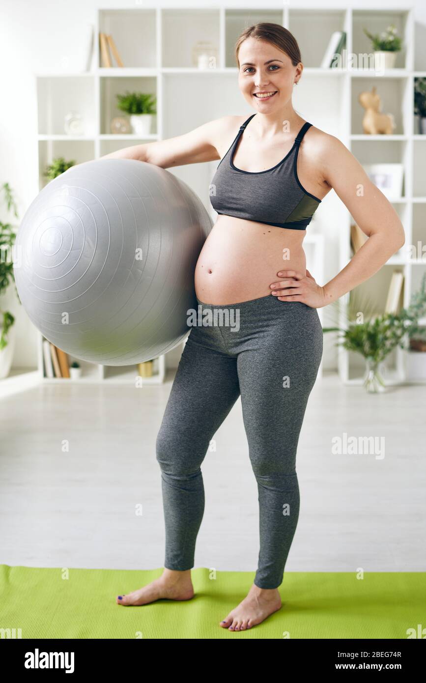 Positive junge schwangere Frau in grauen Leggings und Sport-BH mit Fitball auf Yoga-Matte zu Hause stehen Stockfoto