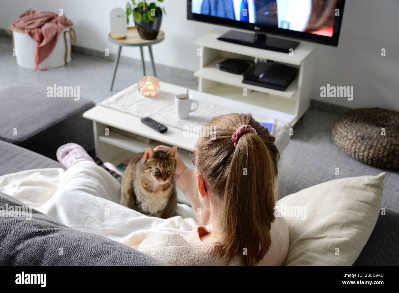 Häusliches Leben mit Haustier zu Hause. Junge Frau sitzt mit ihrer Katze auf der Couch im Wohnzimmer. Sie schaut fern, während sie ihre Katze streichelte. Binge beobachten Stockfoto