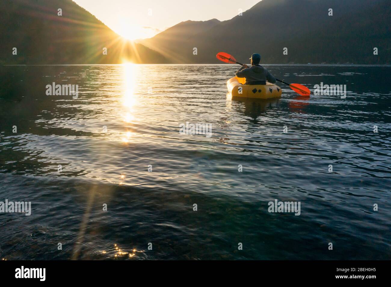 Junger Mann paddelnd auf See bei Sonnenuntergang in Richtung Berge Stockfoto