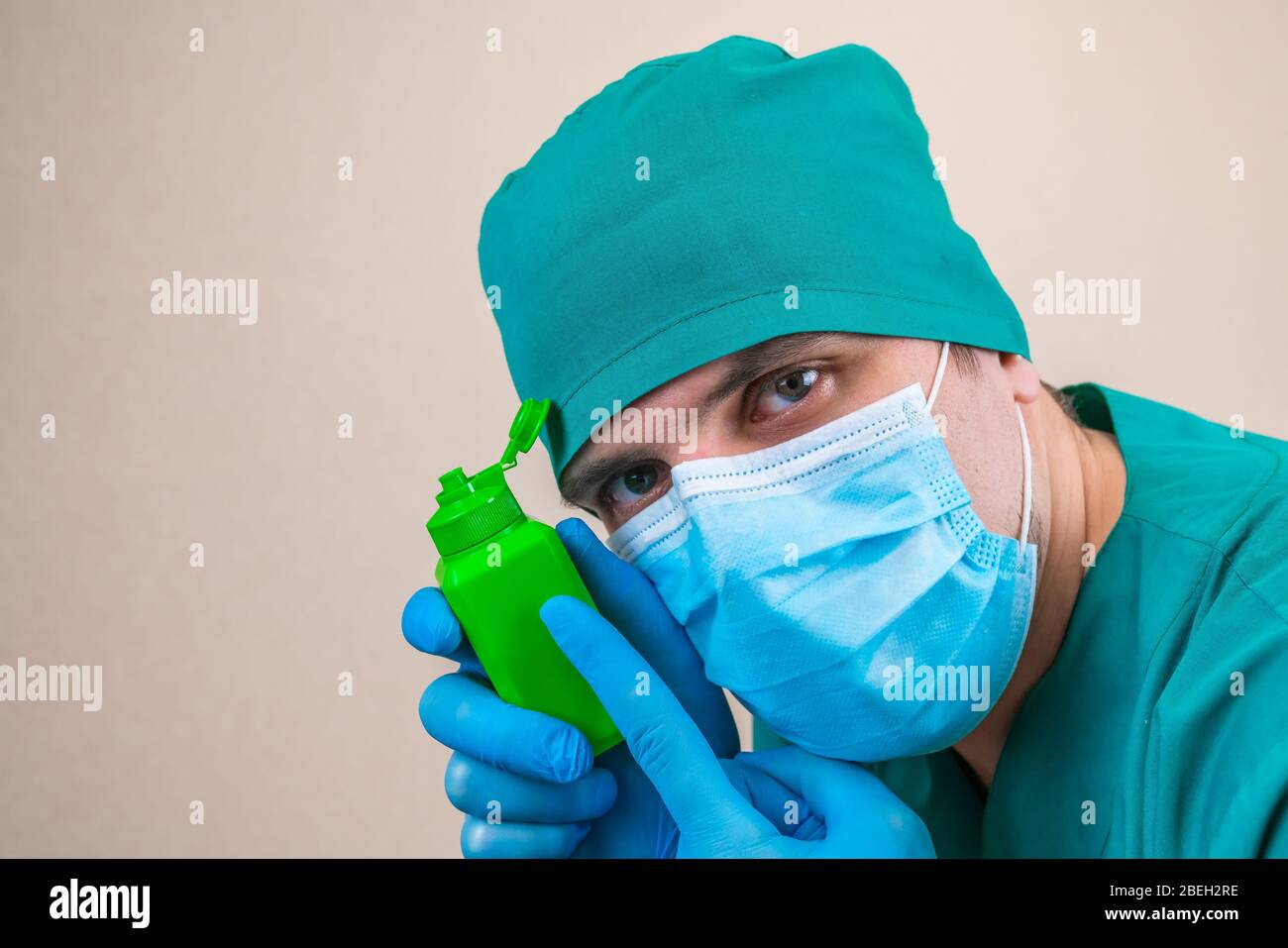 Doktor in grünem Anzug und blauen Maskenhandschuhen mit grüner antiseptischer Flasche in der Hand auf gelbem Hintergrund. Stockfoto