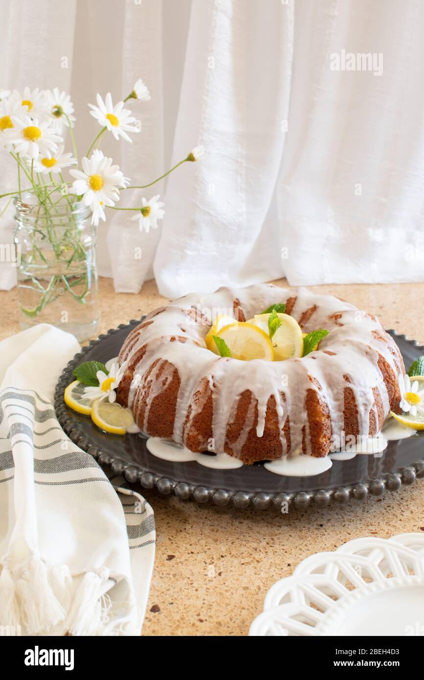 Zitronenkuchen Ricotta mit Glasur auf einem schwarzen dekorativen Teller mit Gänseblümchen in einer Vase im Hintergrund. Stockfoto