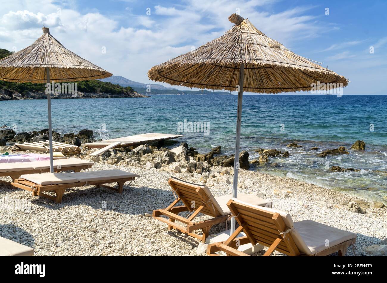 Felsigen Strand am Ufer des Ionischen Meer mit Sonnenliegen und Sonnenschirme Stroh. Blau und tourquse Meer im Hintergrund, niemand. Sommertag. Ksamil, Al Stockfoto