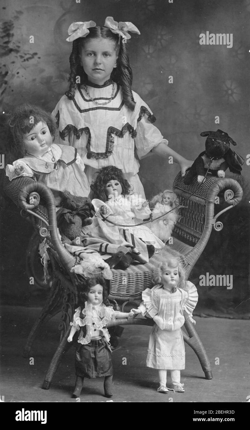 Um 1910 steht ein Mädchen hinter ihrer Puppenfamilie, die auf einem Korbsitz ohne Rücken steht. Das Mädchen ist selbst sehr gut gekleidet in ein ausgefallenes Kleid und zwei Bögen in ihrem langen lockigen Haar. Ihre Familie besteht aus 5 Biskuit-Puppen in verschiedenen Größen und einem Steiff Bear, der in einer großen schwarzen Schleife gekleidet ist. Die große sitzende Puppe hat einen ausgestopften Hund auf Mietvertrag. Der Hund ruht auf dem Stuhlarm. Dieser etwas gefährlich aussehende Hund hat neben dem Halsband auch einen großen schwarzen Bogen um seinen Hals. Um meine anderen Spielzeug-bezogenen vintage Bilder zu sehen, Suche: Prestor vintage Toy [or Kids] Stockfoto