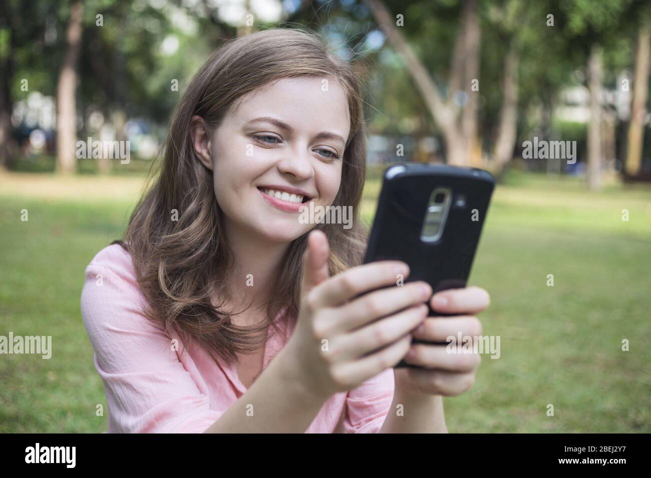 Die junge kaukasische Frau, die lächelnd auf den Bildschirm des Telefons schaut, sieht glücklich aus. Außenbilder. Stockfoto
