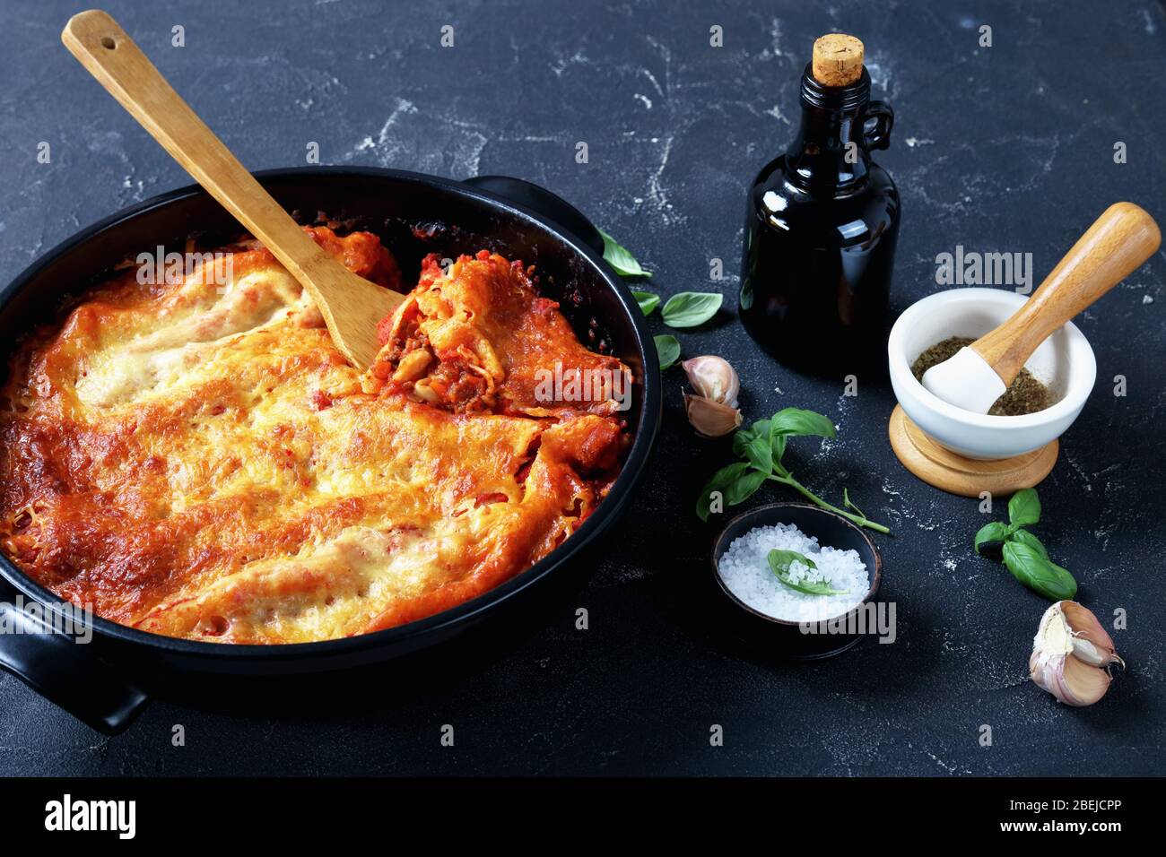 Italienische Cannelloni-Pasta mit Bolognese-Sauce und Mozzarella-Käse auf einer schwarzen Backform auf dunklem Betongrund mit frischem Basilikum gefüllt Stockfoto