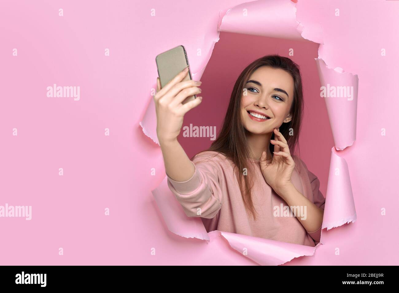 Porträt der schönen lächelnden kaukasischen Frau, die das Smartphone in der Hand hält und selfie auf der vorderen Kamera in zerrissener pinkfarbener Papierwand schießt. Kopierraum Stockfoto