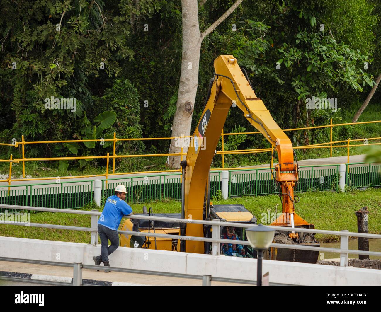 SINGAPUR – 21 FEB 2020 - ein indischer / bangladeschischer Bauarbeiter in Harthut beobachtet einen Bagger bei der Baggerarbeit in einem Naturpark Stockfoto