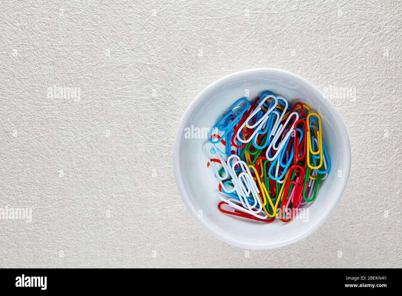 Haufen von bunten Büroklammern in einer weißen Schüssel auf weißem texturiertem Hintergrund.. Makroansicht mit flacher Lay-Ansicht. Stockfoto