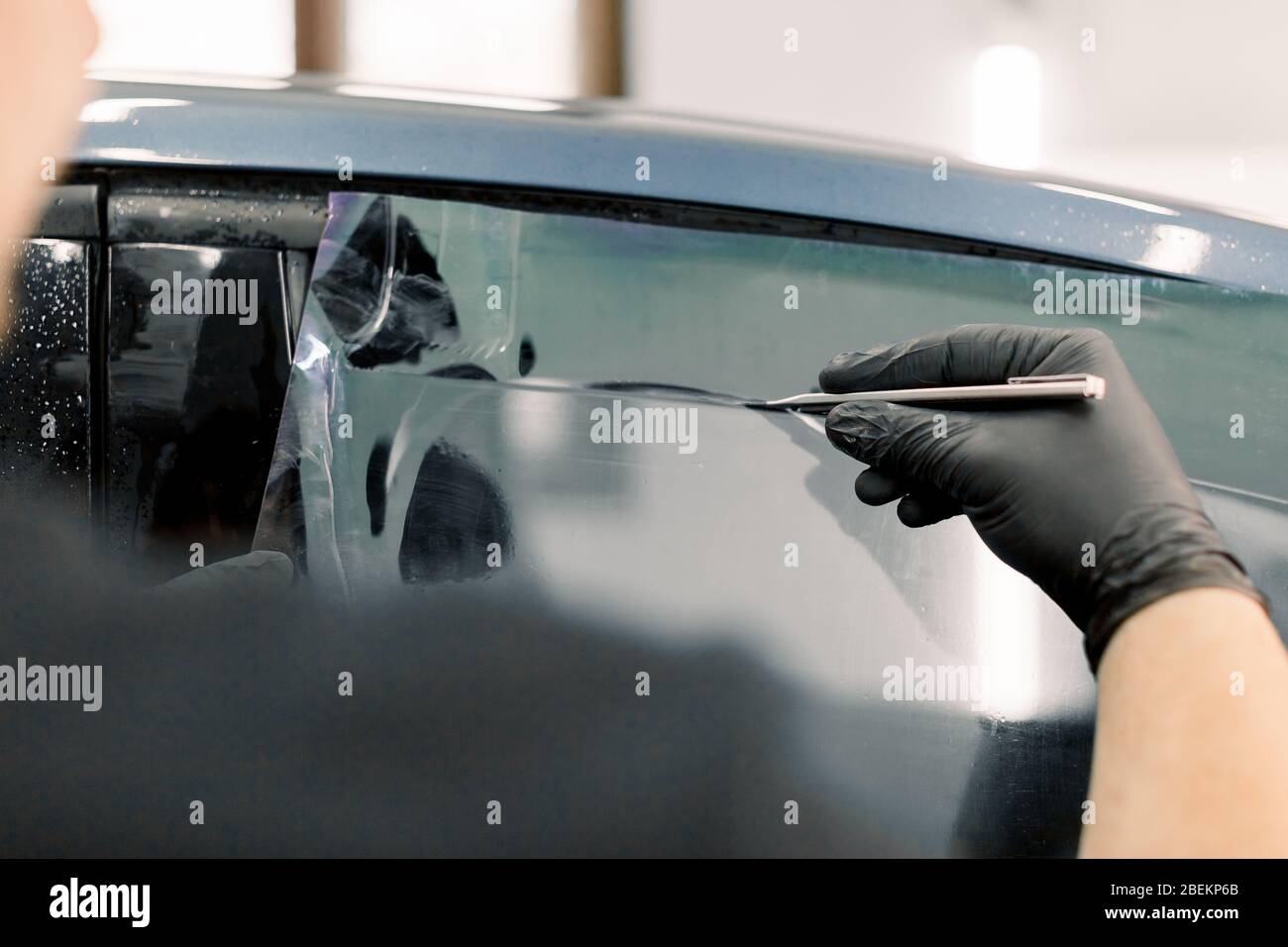 Zugeschnittenes Bild von Händen des Arbeiters in der Garage, die ein Autofenster mit getönter Folie oder Folie färben, die spezielle Klinge oder Messer halten, um den Film zu schneiden. Auto Stockfoto