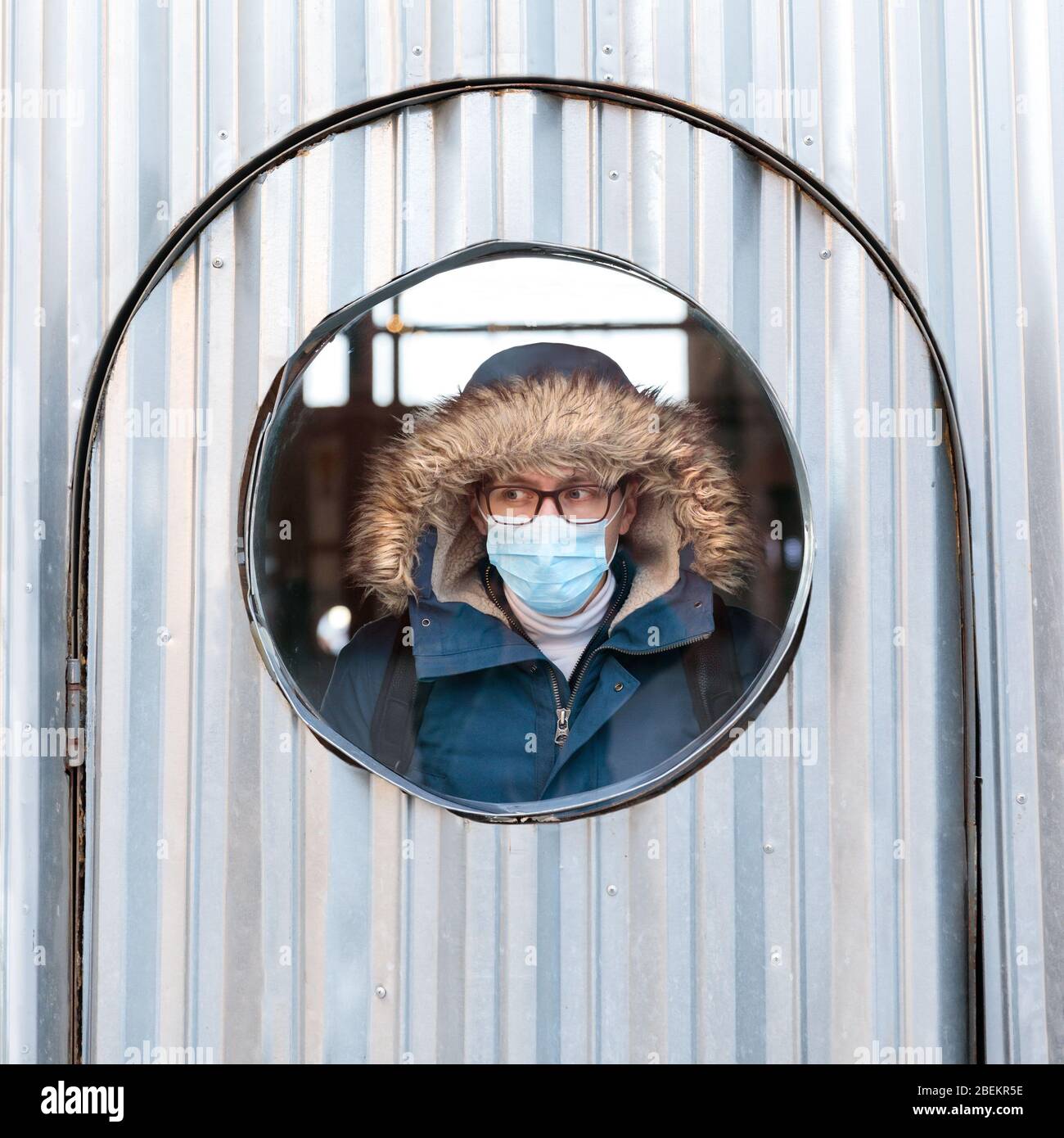 Kranker Mann mit Kapuze blickt aus dem runden Fenster, trägt eine schützende Gesichtsmaske gegen übertragbare Infektionskrankheiten und schützt vor Th Stockfoto Kranker Mann mit Kapuze blickt aus dem runden Fenster, trägt eine schützende Gesichtsmaske gegen übertragbare Infektionskrankheiten und schützt vor Th Stockfoto