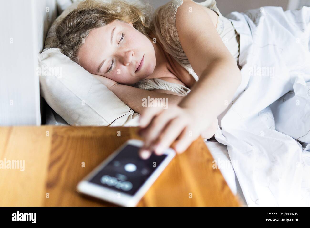 Junge Osteuropäerin, die im Bett liegt und morgens den Wecker auf ihrem Handy abschaltet. Stockfoto