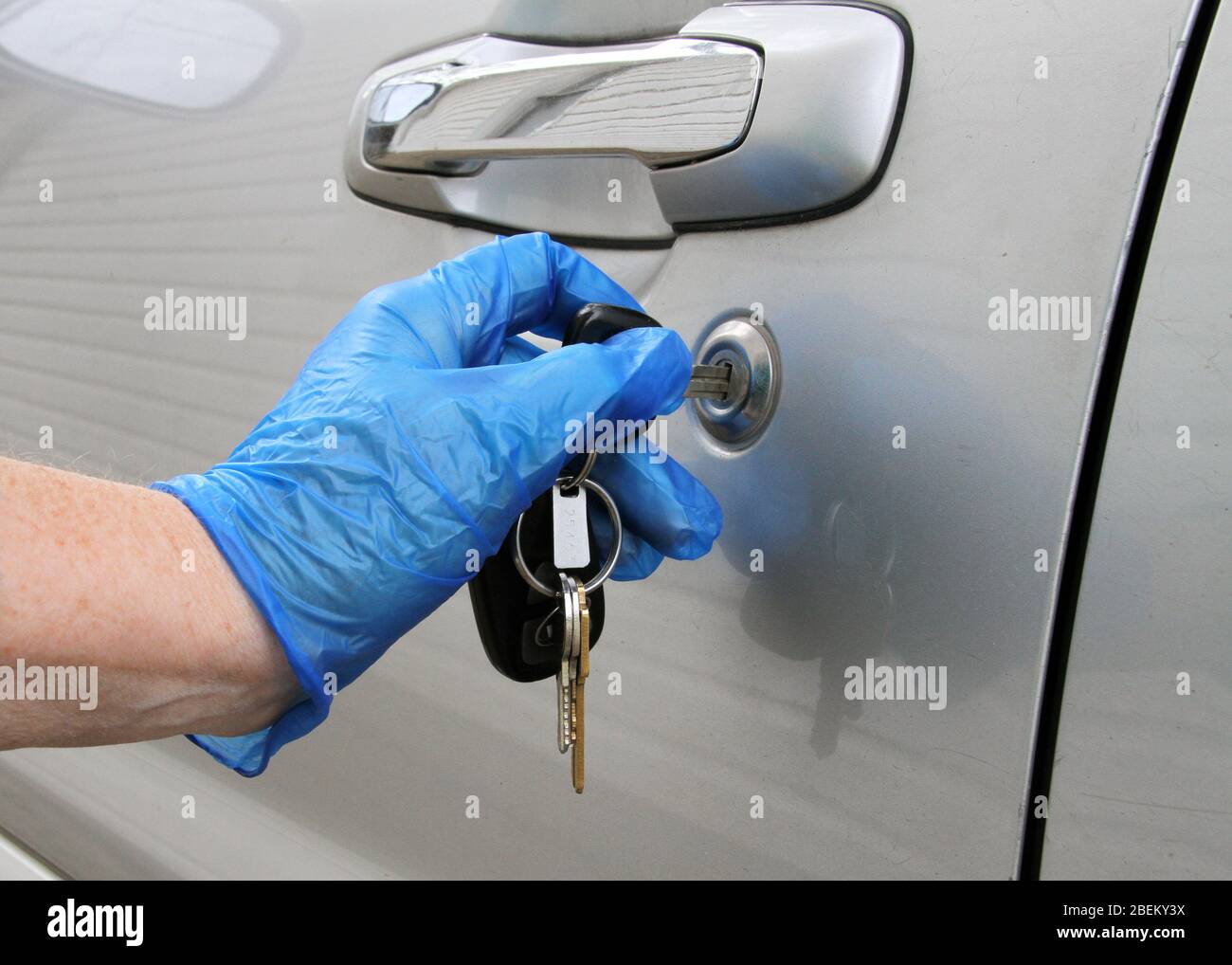 Nahaufnahme einer Hand in einem blauen Latex-Handschuh, der während der Coronavirus-Pandemie eine silberne Autotür mit Schlüsseln entriegelt. Stockfoto