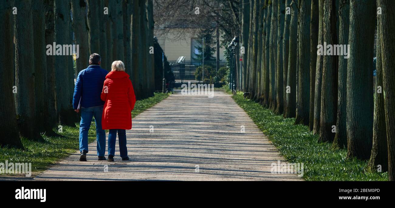 Celle, Deutschland, 27. März 2020: Älteres Paar, das allein auf der Lindenallee im französischen Garten läuft, Frau mit roter Jacke und blonden Haaren Stockfoto