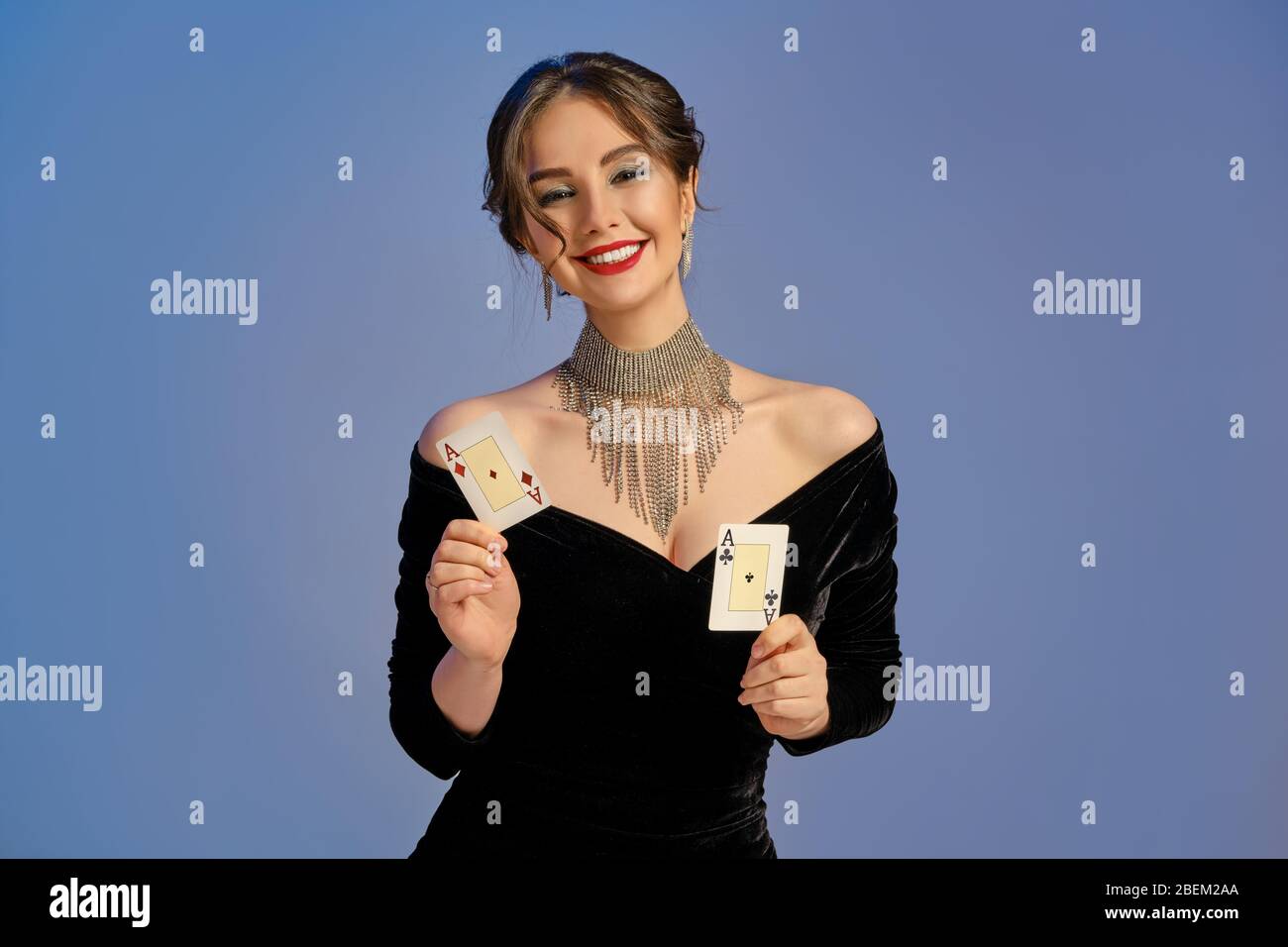 Brunette Mädchen mit nackten Schultern, in schwarzem Kleid und Schmuck.  Lächelnd, zeigt zwei Spielkarten auf blauem Hintergrund. Poker, Casino.  Nahaufnahme Stockfotografie - Alamy