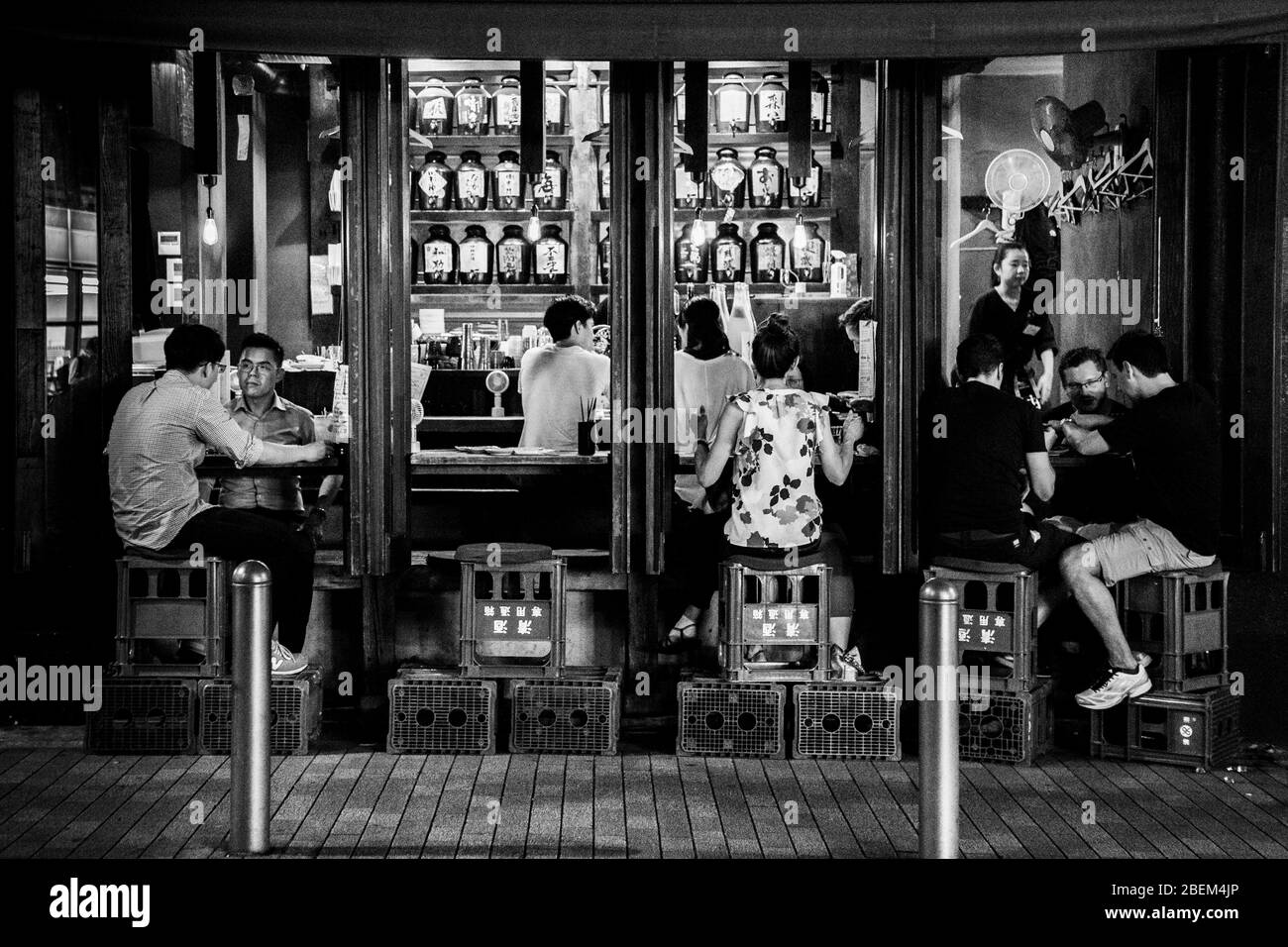 Gäste sitzen auf gestapelten roten Bierkisten vor den offenen Theken einer schmalen Reihe von Bars in Minato, Tokio, Japan. Eine Kellnerin steht lachend in einem Durchgang. Reihen von abgerundeten Sake-Flaschen füllen die Regale in den engen Innenräumen. Stockfoto