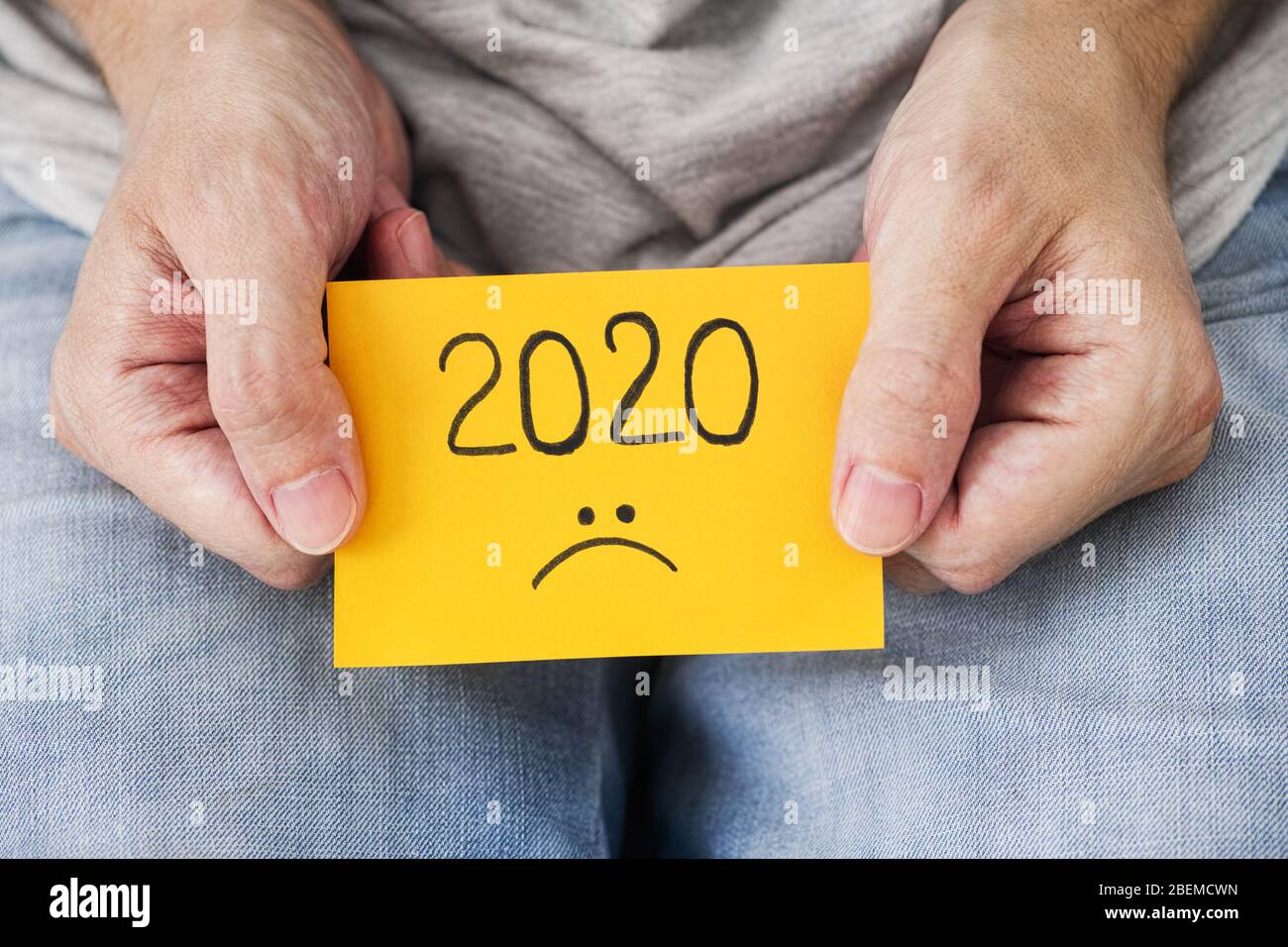Mann, der ein gelbes Stück Papier mit dem Jahr 2020 und einem traurigen Smiley in den Händen hält. 2020 schreckliches Jahr Konzept. Nahaufnahme. Stockfoto
