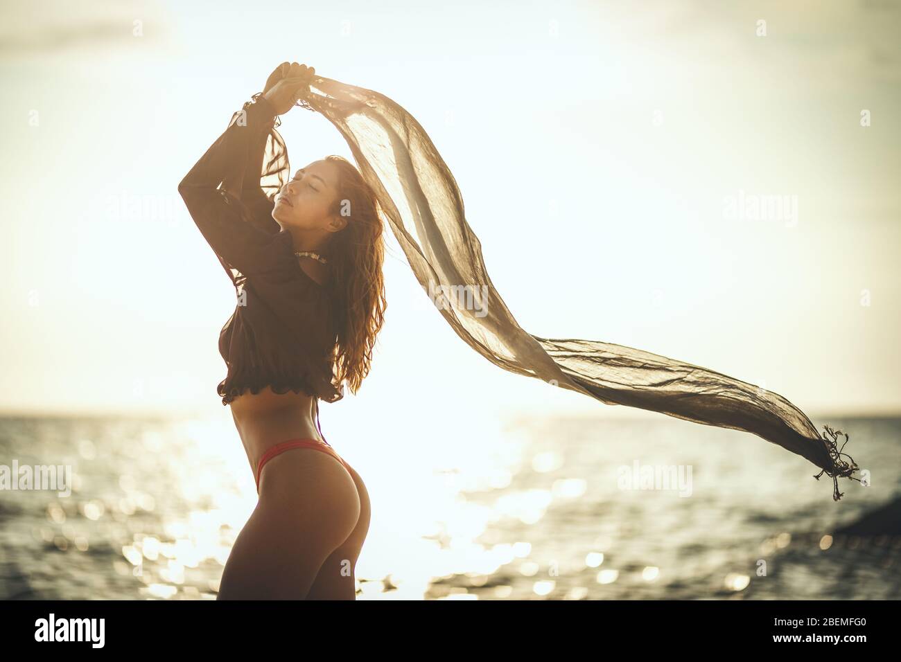 Eine schöne junge Frau hat Spaß und entspannt am Strand bei Sonnenuntergang. Sie posiert und schaut weg mit einem durchsichtigen Schal in den Händen. Stockfoto