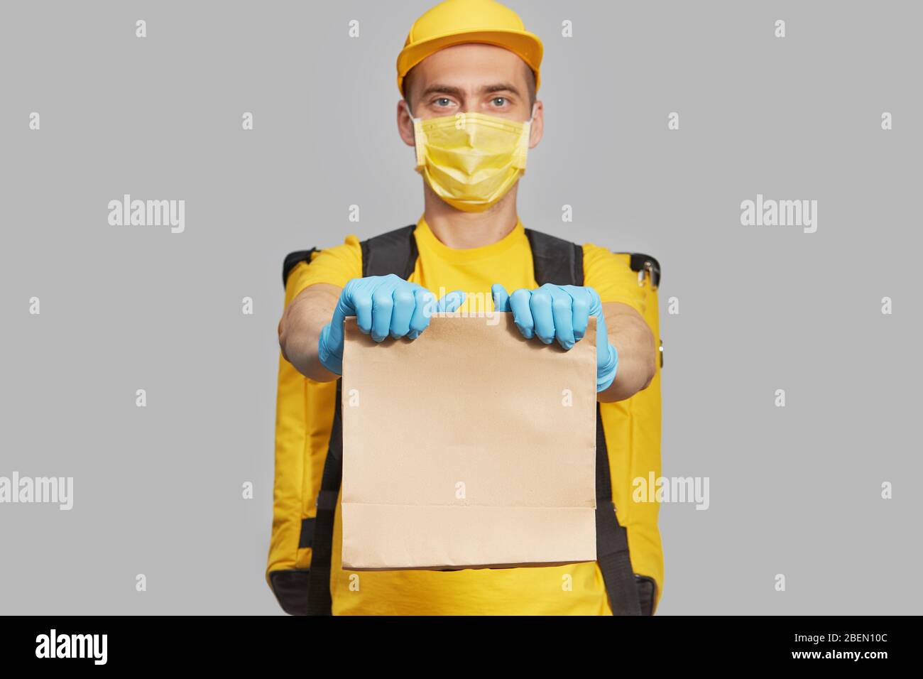 Kurier in gelber Uniform, Schutzmaske und medizinische Handschuhe liefern Essen und Kaffee zum Mitnehmen. Lieferservice unter Quarantäne. Kopieren Sie den Bereich für den tex Stockfoto