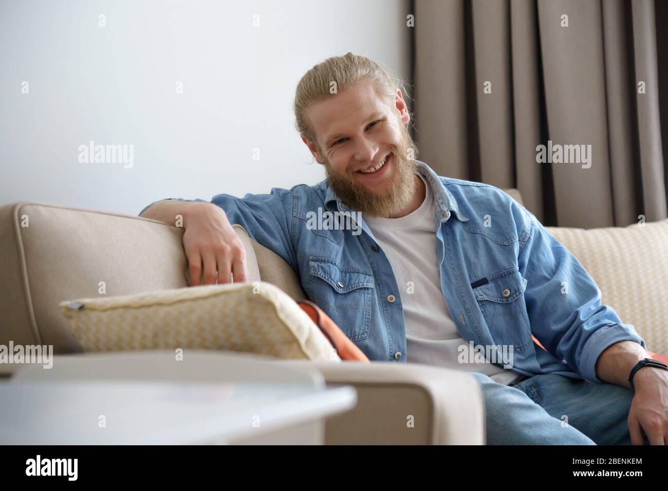 Lächelnder junger Mann, der sich zu Hause auf dem Sofa entspannt, Kamera, Porträt anschaut. Stockfoto