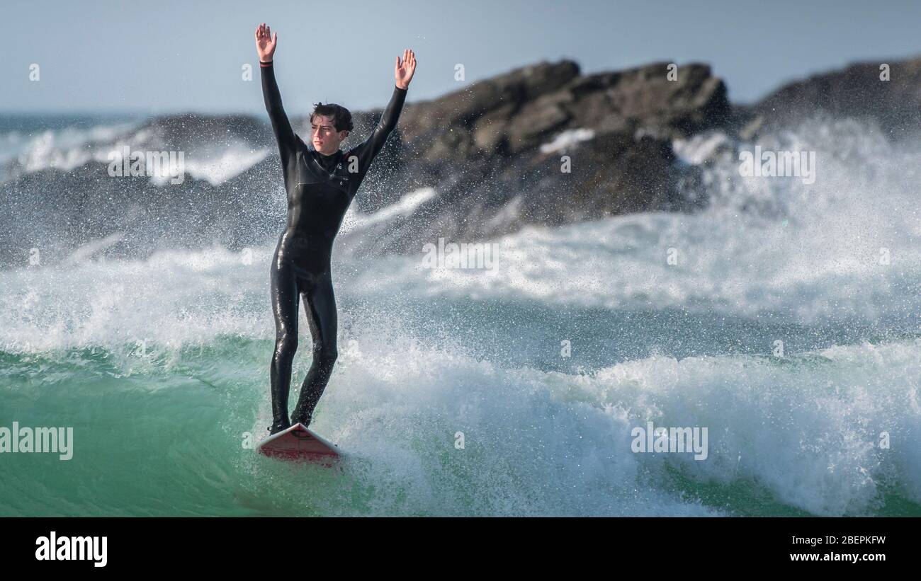 Ein Panoramabild eines jungen Surfers, der seinen Freunden winkt, während er eine Welle bei Fistral in Newquay in Cornwall reitet. Stockfoto