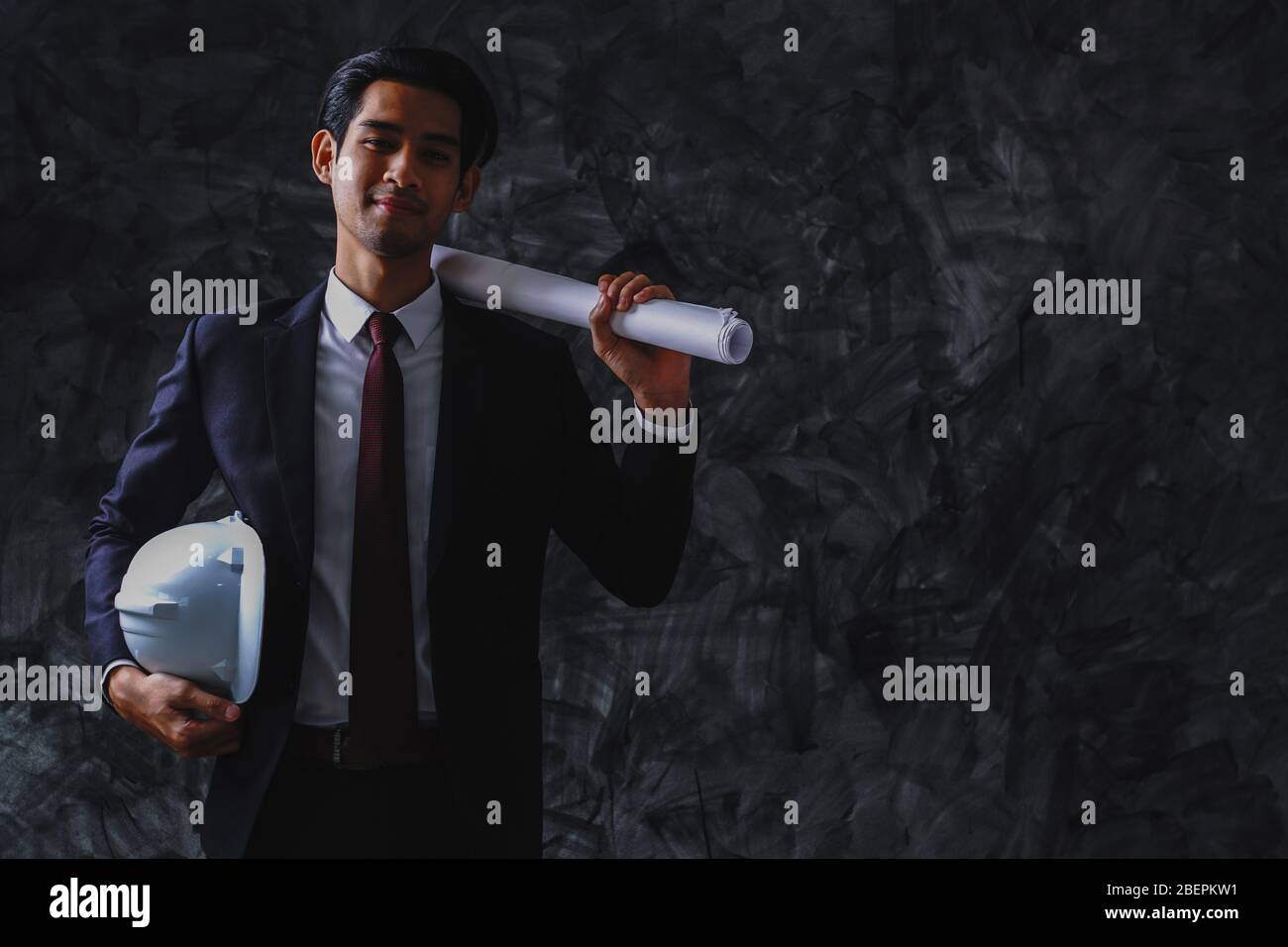 Junge Wirtschaftsingenieurwesen mit weißem Helm und blauem Druck auf dunklem Hintergrund, smart und zuversichtlich Arbeiter. Stockfoto