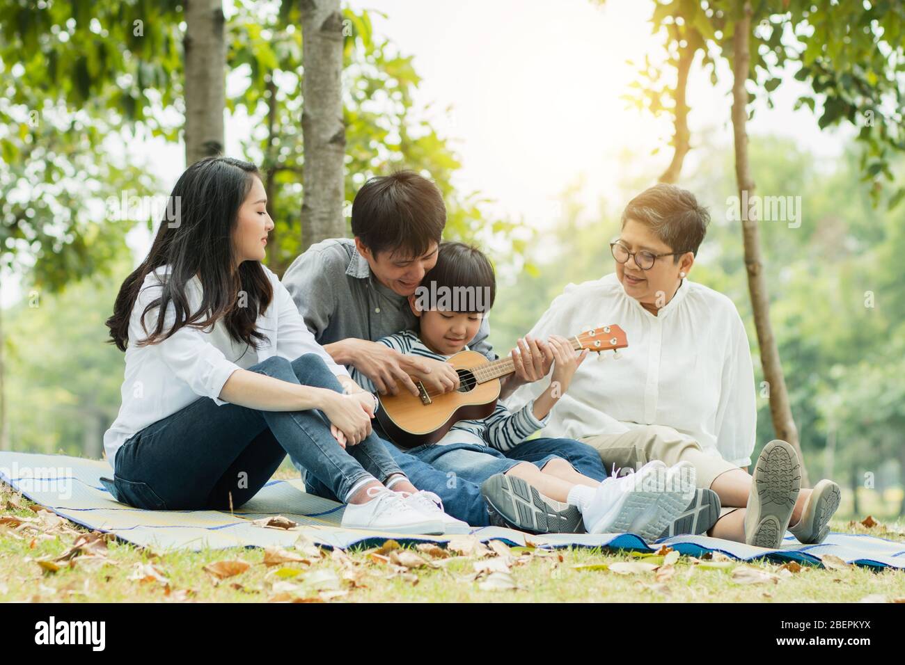 Glückliche Familie mit Oma, Mama mit Papa, der Sohn Gitarre spielt und ein Lied im Park singt, genießen und entspannen Leute Picknick draußen Stockfoto
