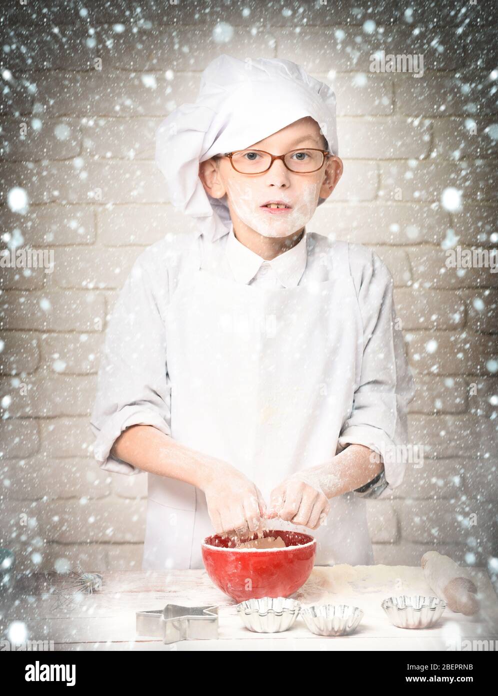 Junge kleine süße Koch Koch Koch in weißer Uniform und Hut auf glückliches Gesicht mit Gläsern stehen in der Nähe Tisch mit Rollen-PIN Formen für Kuchen rote Schüssel, und das Spielen mit Mehl auf Backstein Wand Hintergrund Stockfoto