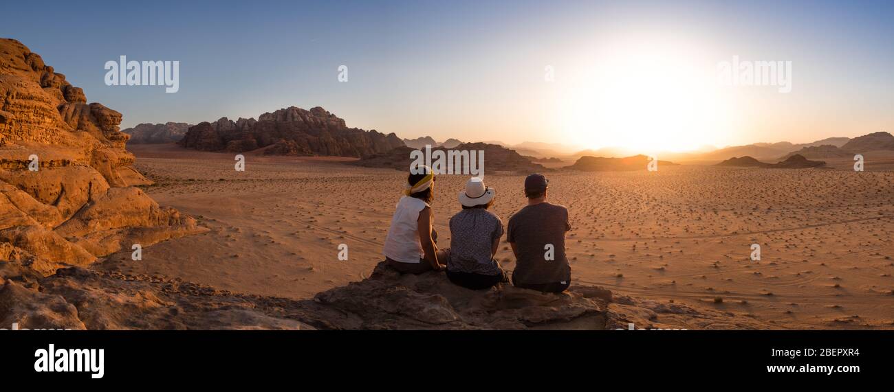 Drei Touristen sitzen auf einer felsigen Plattform und beobachten den Sonnenuntergang in Wadi Rum, Petra Stockfoto