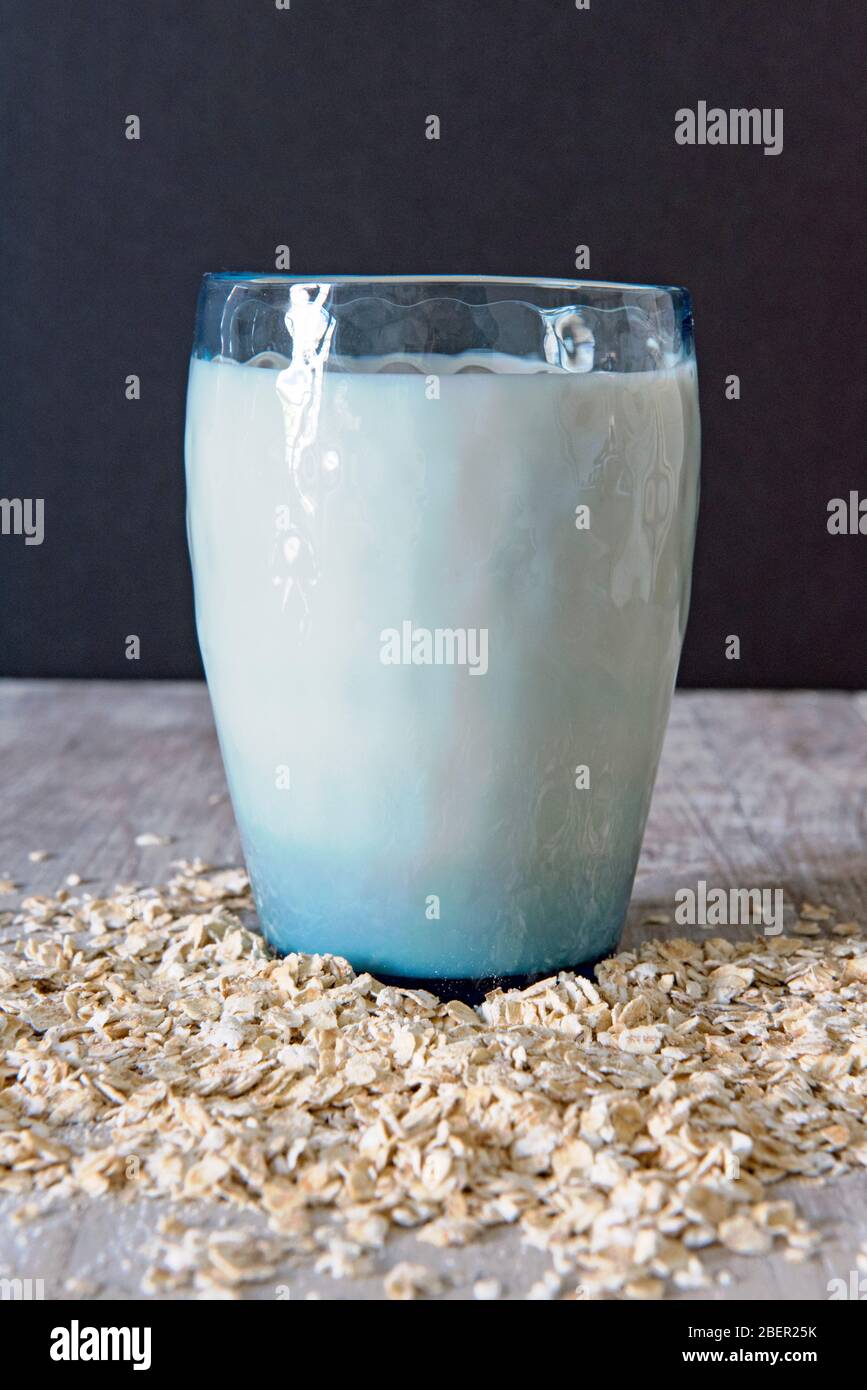 Blaues Vintage Glas voller veganer Hafermilch oder Getränk mit Hafer, der ringsherum gestreut ist und schwarzem Hintergrund Stockfoto