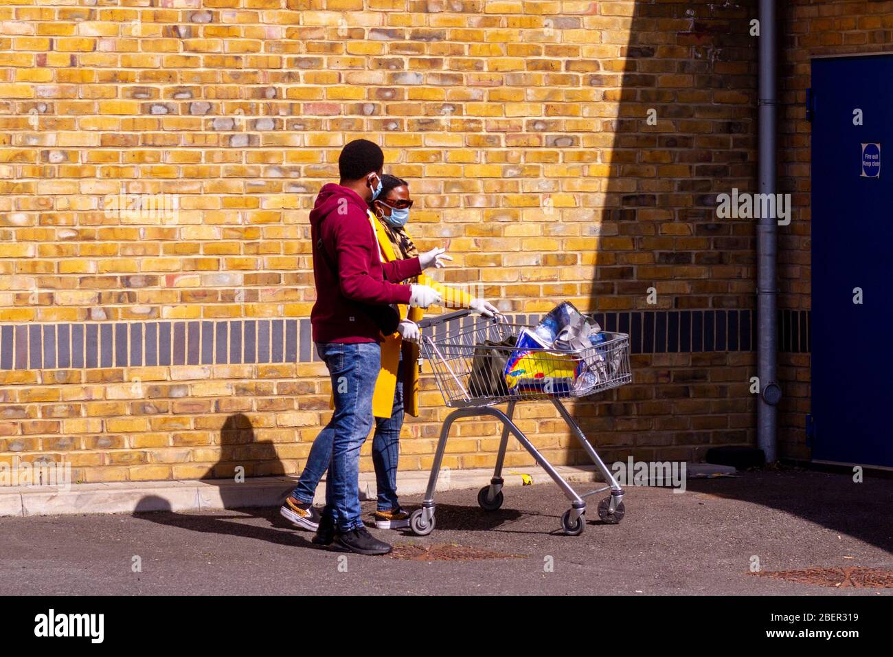 Homerton, London, Großbritannien, 7. April 2020: Zwei Menschen warten mit Trolley und Einkaufstaschen sowie Gesichtsmasken und Handschuhen in der Schlange Stockfoto