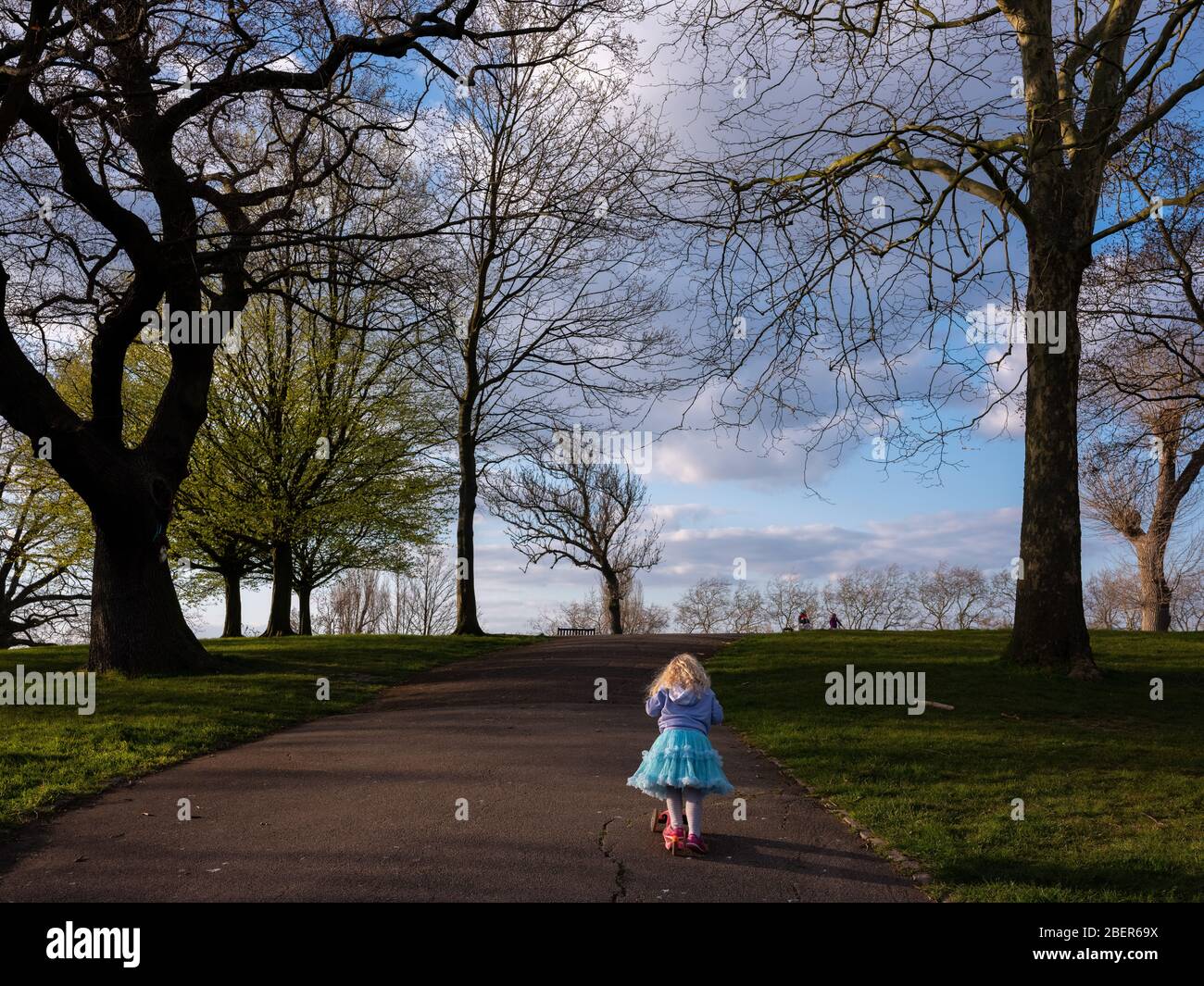 Ein junges Mädchen von 2 (fast 3), das alleine in einem Londoner Park einen Hügel hinaufscooting. Roundwood Park, London. Stockfoto