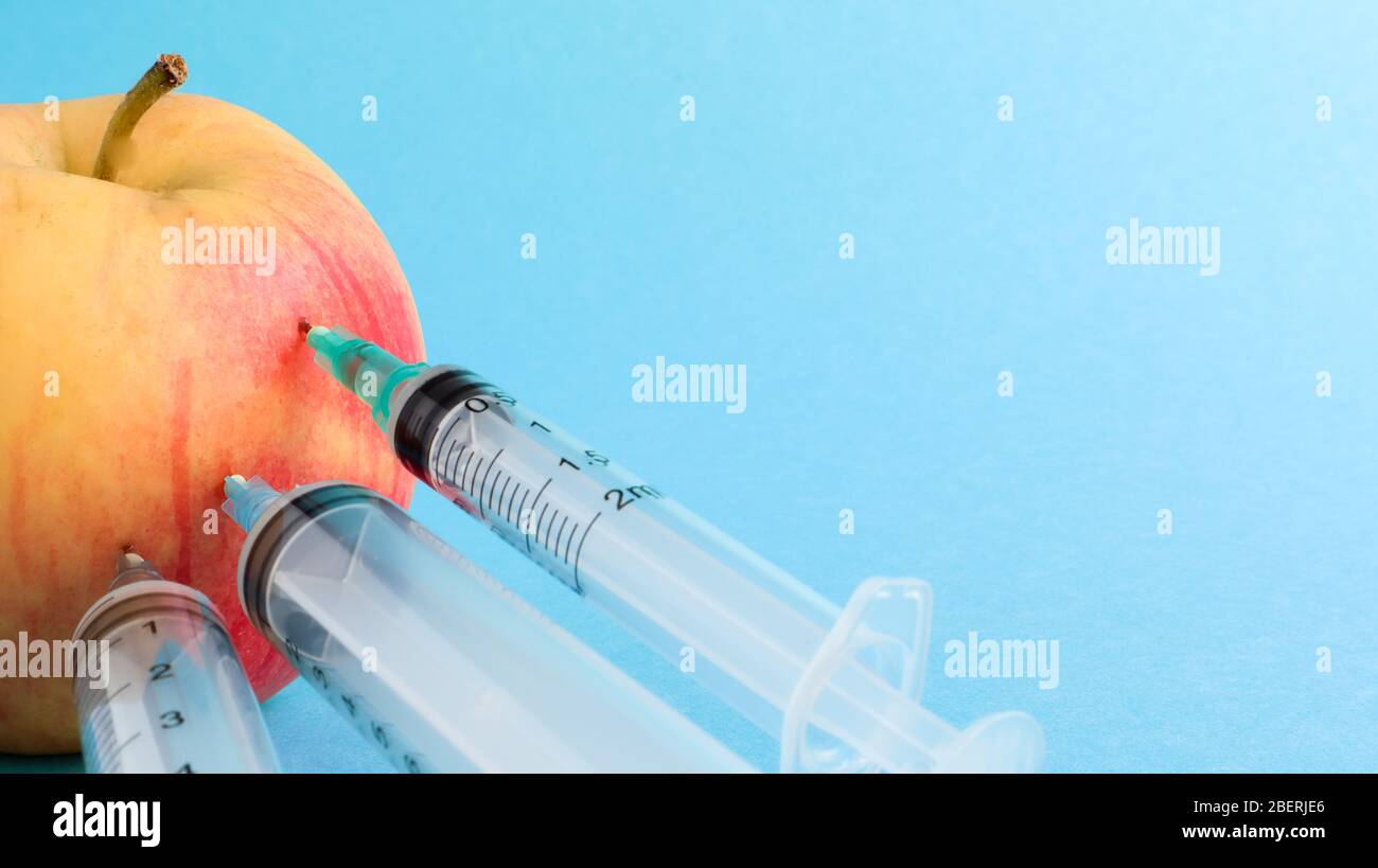 Drei verschiedene medizinische Spritzen, die tief in einen Apfel auf blauem Hintergrund stecken. Die Frucht ist mit Spritzen übersät. Ein reifer roter Apfel, der von al durchbohrt wurde Stockfoto