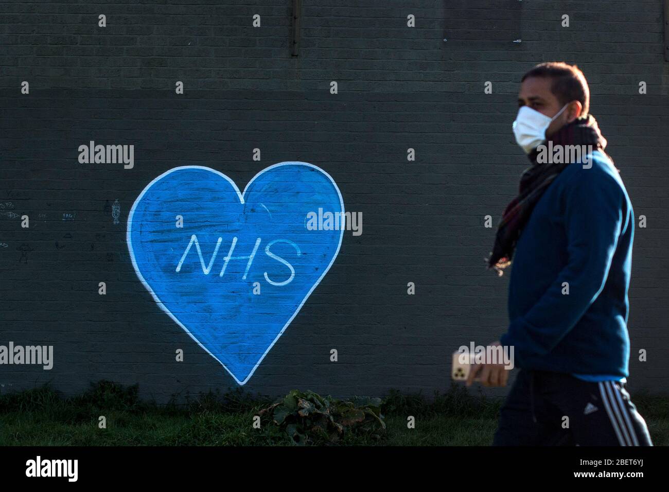 Ein Mann in einer Gesichtsmaske geht an Graffiti vorbei, um das NHS auf Wanstead Flats in East London zu unterstützen, während Großbritannien weiterhin in der Lockdown-Richtung arbeitet, um die Ausbreitung des Coronavirus einzudämmen. Stockfoto